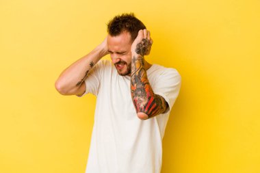 Genç dövmeli beyaz adam sarı arka planda izole edilmiş kulakları elleriyle kapatıyor..