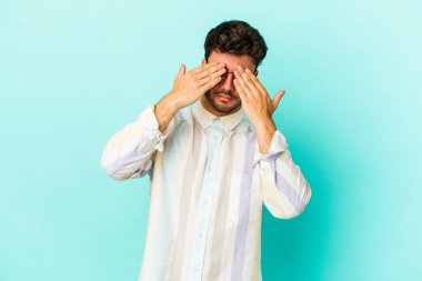 Mavi arka planda izole edilmiş genç beyaz adam elleriyle gözlerini kapatmaktan korkuyor..
