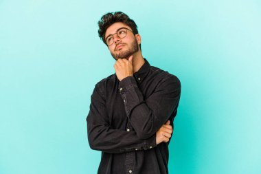 Kafkasyalı genç adam mavi arka planda izole edilmiş düşünüyor ve bakıyor, yansıtıyor, düşünüyor, bir fantezi kuruyor..
