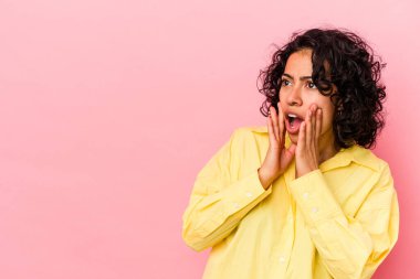 Genç kıvırcık Latin kadın pembe arka planda izole edilmiş. Gördüğü bir şey yüzünden şoka uğruyor..