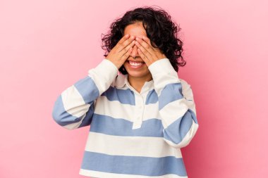 Pembe arka planda soyutlanmış genç kıvırcık Latin kadın elleriyle gözlerini kapatmaktan korkuyor..