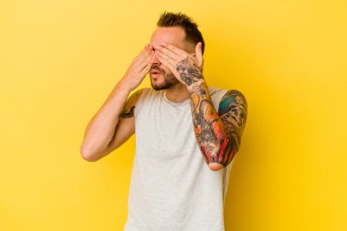 Sarı arka planda izole edilmiş genç dövmeli beyaz adam elleriyle gözlerini kapatmaktan korkuyor..