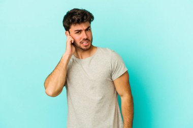 Mavi arka planda izole edilmiş genç beyaz adam kulaklarını elleriyle kapatıyor..