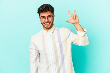 Mavi arka planda izole edilmiş genç beyaz adam parmaklarıyla kaya hareketi gösteriyor.