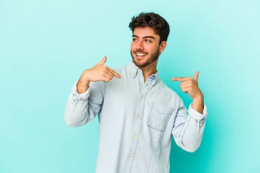 Mavi arka planda izole edilmiş genç beyaz adam parmağını göstererek şaşırdı, geniş bir gülümsemeyle..