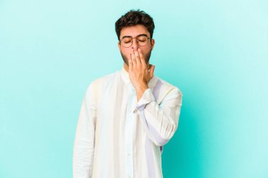 Mavi arka planda izole edilmiş genç beyaz adam esniyor. Ağzı eliyle kapatan yorgun bir hareket sergiliyor..