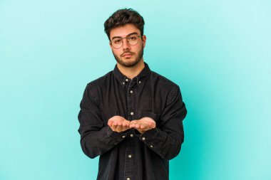 Kafkasyalı genç adam mavi arka planda izole edilmiş. Elinde avuç içi olan bir şey var ve kameraya sunuyor..