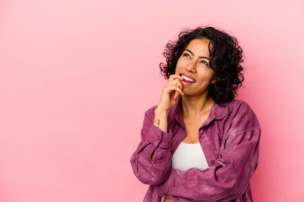 Young curly latin woman isolated on pink background relaxed thinking about something looking at a copy space.
