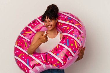 Beyaz arka planda izole edilmiş şişme bir donut tutan genç melez kadın sanki seni davet ediyormuş gibi seni işaret ediyor..