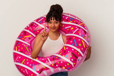 Beyaz arka planda izole edilmiş şişirilebilir donut tutan genç bir ırk kadını parmak izleriyle bir numarayı gösteriyor..