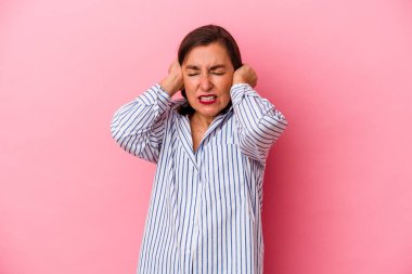 Orta yaşlı beyaz kadın pembe arka planda izole edilmiş kulakları elleriyle kapatıyor..