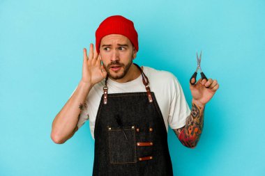 Genç dövmeli berber mavi arka planda izole edilmiş bir şekilde bir dedikoduyu dinlemeye çalışıyor..