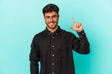 Kafkasyalı genç adam mavi arka planda izole edilmiş. Elinde işaret parmaklarıyla küçük bir şey tutuyor, gülümsüyor ve kendinden emin..