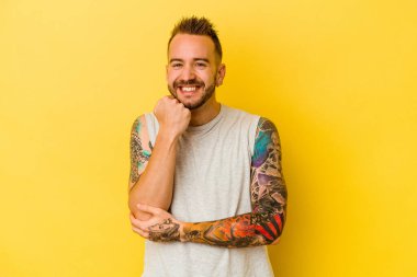 Sarı arka planda izole edilmiş genç dövmeli beyaz adam mutlu ve kendinden emin gülümsüyor, çenesine elleriyle dokunuyor..