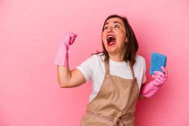 Orta yaşlı beyaz kadın evini temizliyor. Pembe arka planda izole edilmiş. Zaferden sonra yumruğunu kaldırıyor..