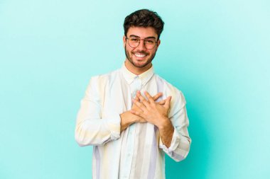 Kafkasyalı genç adam mavi arka planda izole olmuş gülüyor, elini yüreğine koyuyor, mutluluk kavramını koruyor..