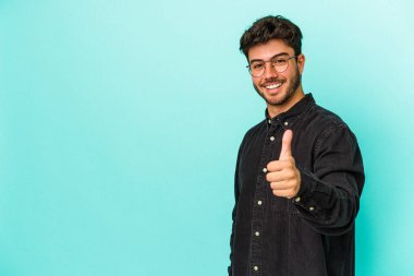Mavi arka planda izole edilmiş genç beyaz adam gülümsüyor ve başparmağını kaldırıyor.