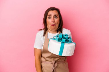 Orta yaşlı pasta şefi kadın mavi arka planda izole edilmiş bir pasta tutuyor omuzlarını silkiyor ve gözleri açık..