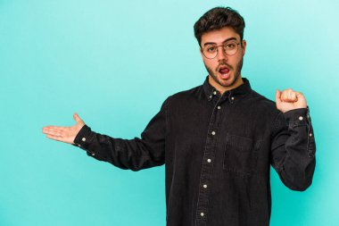 Mavi arka planda izole edilmiş genç beyaz bir adam avucunun içinde fotokopi alanı tutuyor, el üstünde tutuyor. Şaşırmış ve mutlu..