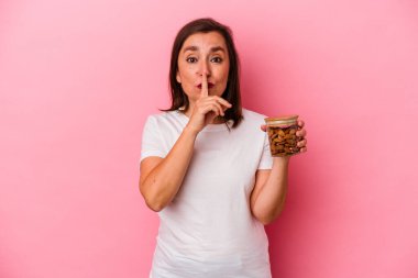 Orta yaşlı beyaz bir kadın pembe arka planda izole edilmiş bir badem kavanozunu tutuyor ya sır saklıyor ya da sessizlik istiyor..