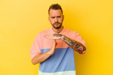 Sarı arka planda izole edilmiş genç dövmeli beyaz adam bir mola hareketi gösteriyor..