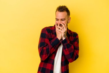 Sarı arka planda izole edilmiş genç dövmeli beyaz adam bir şeye gülüyor, elleri ağızda..