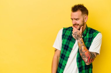 Genç dövmeli beyaz adam sarı arka planda izole edilmiş kafasının arkasına dokunuyor, düşünüyor ve bir seçim yapıyor..