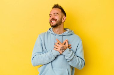Sarı arka planda izole edilmiş genç, dövmeli beyaz adam gülüyor, ellerini kalbinde tutuyor, mutluluk kavramını koruyor..