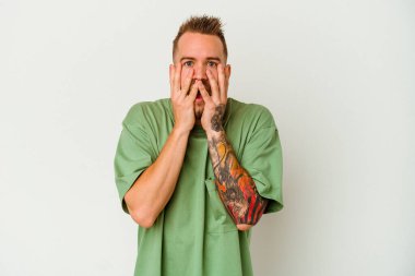 Beyaz arka planda izole edilmiş genç, dövmeli beyaz adam şok olmuş, ağzı ellerle kaplanmış, yeni bir şey keşfetmeye hevesli..