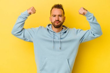 Sarı arka planda izole edilmiş genç dövmeli beyaz adam kadınsı gücün sembolü olan kollarıyla güçlü bir hareket gösteriyor.