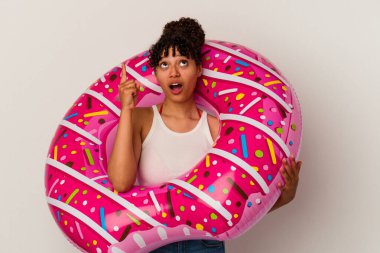 Beyaz arka planda izole edilmiş şişirilebilir hava donutu tutan genç bir ırk kadını ağzı açık bir şekilde yukarı işaret ediyor..