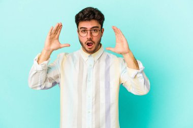 Mavi arka planda izole edilmiş genç beyaz bir adam zaferi ya da başarıyı kutluyor, şaşırıyor ve şok oluyor..