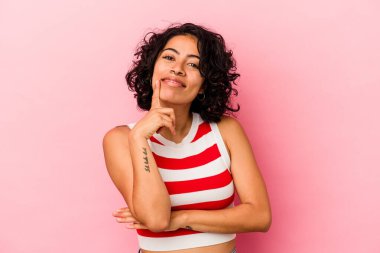 Genç kıvırcık Latin kadın pembe arka planda izole edilmiş mutlu ve kendinden emin gülümsüyor, elleriyle çenesine dokunuyor..