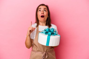 Orta yaşlı pasta şefi kadın elinde mavi arka planda izole edilmiş bir pasta tutuyor ve ağzı açık bir şekilde üst tarafı gösteriyor..