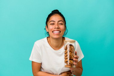 Mavi arka planda izole edilmiş kurabiye kavanozunu tutan genç Latin kadın gülüyor ve eğleniyor..