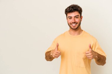 Beyaz arka planda izole edilmiş genç beyaz adam gülümsüyor ve başparmağını kaldırıyor.