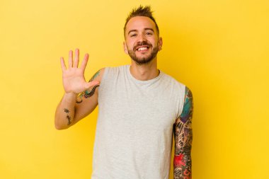 Sarı arka planda izole edilmiş genç dövmeli beyaz adam gülümseyerek beş numarayı parmaklarıyla gösteriyor..
