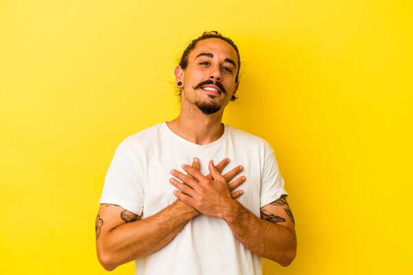 Young caucasian man with long hair isolated on yellow background has friendly expression, pressing palm to chest. Love concept.
