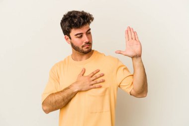 Beyaz arka planda izole edilmiş genç beyaz adam yemin ediyor, elini göğsüne koyuyor..