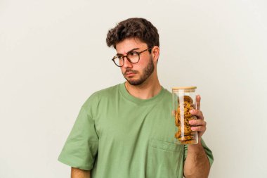 Beyaz arka planda izole edilmiş kurabiye kavanozu tutan Kafkasyalı genç adam kafası karışmış ve kendinden emin değil..