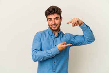 Beyaz arka planda izole edilmiş genç beyaz bir adam işaret parmaklarıyla küçük bir şey tutuyor, gülümsüyor ve kendinden emin..