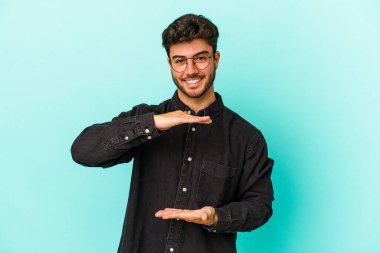 Mavi arka planda izole genç beyaz adam iki eliyle bir şey tutuyor, ürün sunumu..