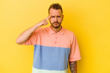 Sarı arka planda izole edilmiş genç dövmeli beyaz adam şakağı parmağıyla işaret ediyor, düşünüyor ve bir göreve odaklanıyor..