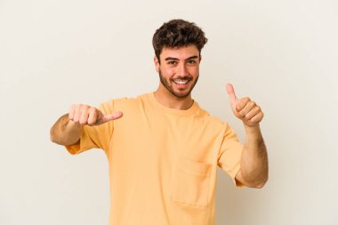 Beyaz arka planda izole edilmiş genç beyaz bir adam iki parmağını da kaldırıyor, gülümsüyor ve kendinden emin..