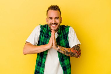 Sarı arka planda izole edilmiş genç, dövmeli beyaz bir adam el ele tutuşup ağız kenarında dua ediyor, kendinden emin hissediyor..
