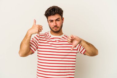 Beyaz arka planda izole edilmiş genç beyaz bir adam baş parmaklarını aşağı ve yukarı kaldırıyor, zor seçim konsepti