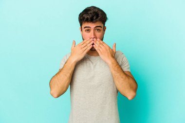 Mavi arka planda izole edilmiş genç beyaz adam elleriyle ağzını kapatırken şok oldu..