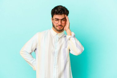 Kafkasyalı genç adam mavi arka planda izole edilmiş. Yorgun ve uykulu. Elini başının üstünde tutuyor..