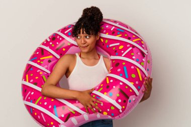 Beyaz arka planda izole edilmiş şişme bir donut tutan genç melez kadın şaşkın ve kararsız hissediyor..