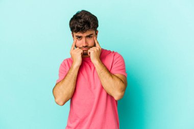 Kafkasyalı genç adam mavi arka planda izole edilmiş, ağlıyor, bir şeyden memnun değil, acı ve kafa karışıklığı konsepti..